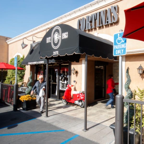 Exterior of Cortina's Italian Market & Pizzeria with a black awning, red scooter, and people entering.