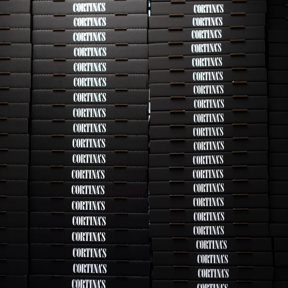 Stacks of black Cortina's pizza boxes with white lettering, ready for delivery.