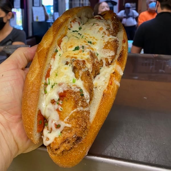 Hand holding a chicken parmesan sub sandwich with melted cheese and red sauce at Cortina's.