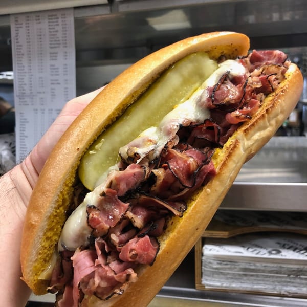 A hand holds a Cortina's Italian Market & Pizzeria pastrami sandwich with melted cheese and a pickle.