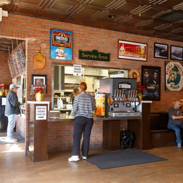 Customers at Cortina's Italian Market & Pizzeria, with a service counter, soda fountain, and brick walls.