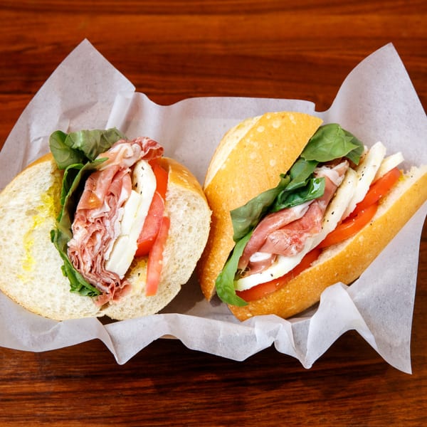 Two Italian sandwiches with prosciutto, mozzarella, tomatoes, and basil on a wooden table.