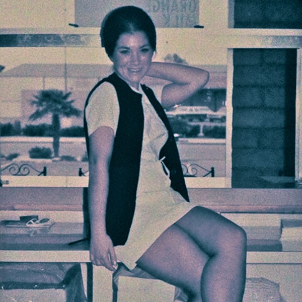 A smiling woman in a white dress and dark vest sits indoors, with a palm tree visible outside.