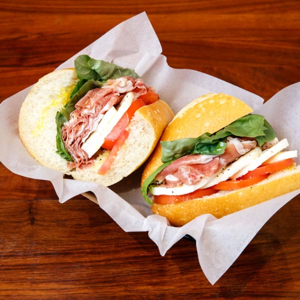 Two Italian sandwiches with prosciutto, mozzarella, tomatoes, and basil on a wooden table.