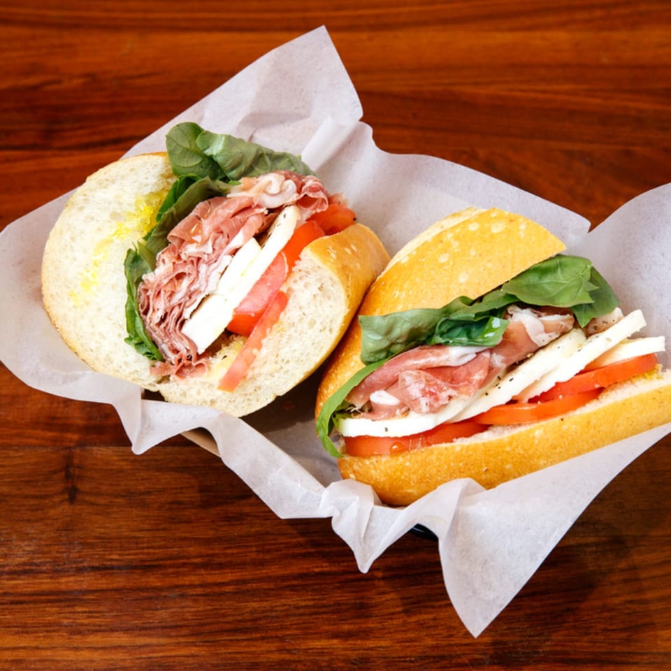 Two Italian sandwiches with prosciutto, mozzarella, tomatoes, and basil on a wooden table.