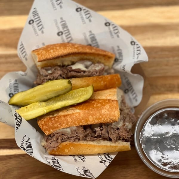 Two Italian beef sandwiches with melted cheese and pickles, served with au jus, on Cortina's branded paper.