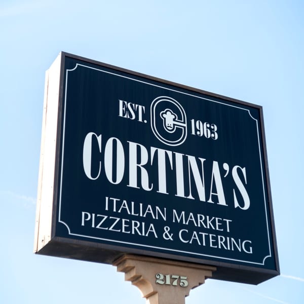 Cortina's Italian Market & Pizzeria sign, black with white lettering, against a clear blue sky.