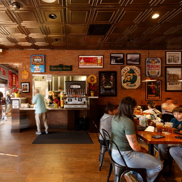 Diners seated at tables in Cortina's Italian Market & Pizzeria, with a service counter and brick walls.