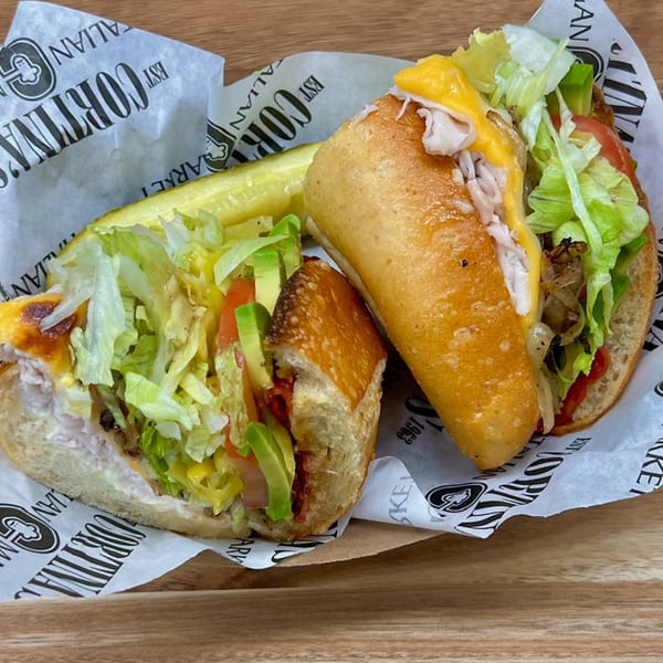 Two Italian subs with turkey, cheese, lettuce, tomato, avocado, and a pickle spear.