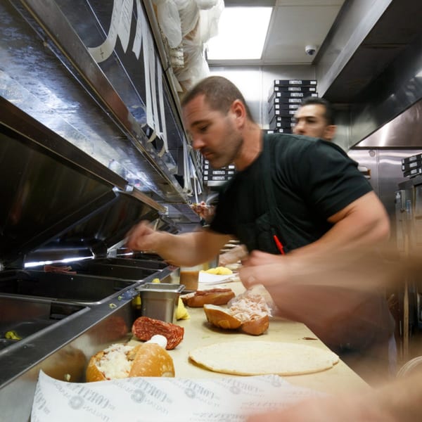 Cortina's Italian Market & Pizzeria chef preparing sandwiches and pizza in a busy kitchen.