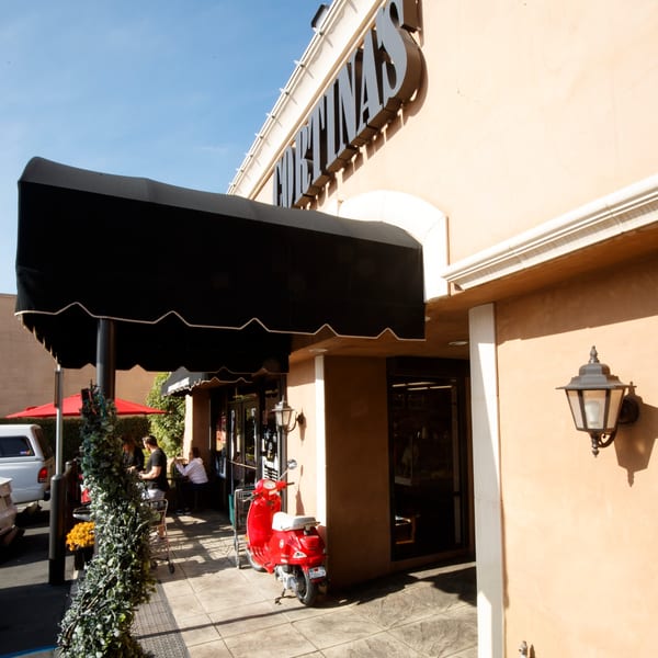 Cortina's Italian Market & Pizzeria exterior with a black awning, red scooter, and outdoor seating.