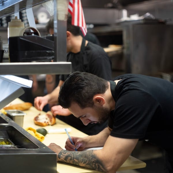 Cortina's Italian Market & Pizzeria chefs preparing food and writing orders in a busy kitchen.