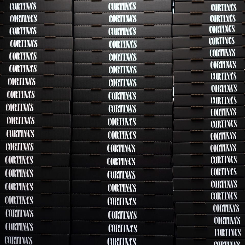 Stacks of black pizza boxes with "CORTINA'S" printed in white on each, ready for delivery.