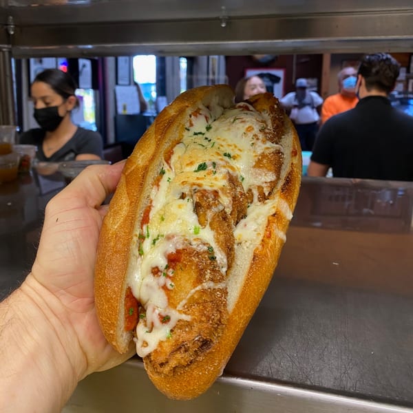 A hand holds a cheesy chicken parmesan sub with marinara sauce, with people in the background.