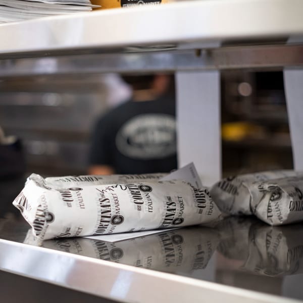 Cortina's Italian Market sandwiches, wrapped in branded paper, sit on a reflective counter.