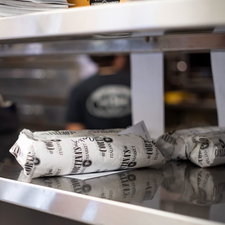 Cortina's Italian Market sandwiches, wrapped in branded paper, sit on a reflective counter.