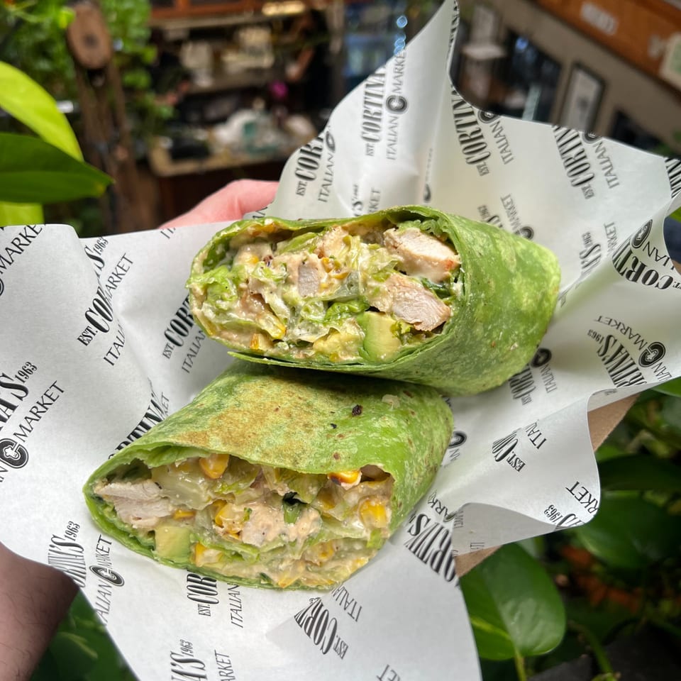 Two green wraps filled with chicken, lettuce, corn, avocado, and creamy sauce on Italian-themed wrapping paper.