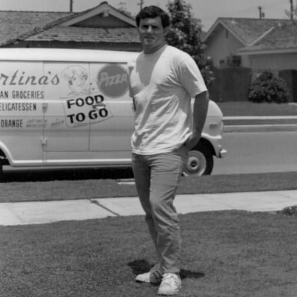 A man stands in front of a Cortina's Italian Market & Pizzeria delivery van in black and white.