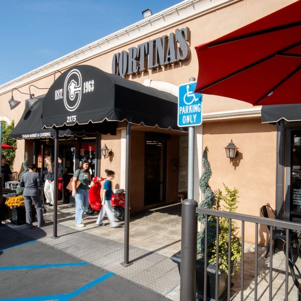 The exterior of Cortina's Italian Market & Pizzeria, with a black awning and people entering.