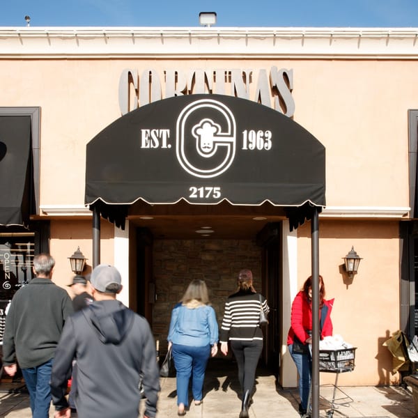 People entering Cortina's Italian Market & Pizzeria under a black awning with the restaurant's logo.