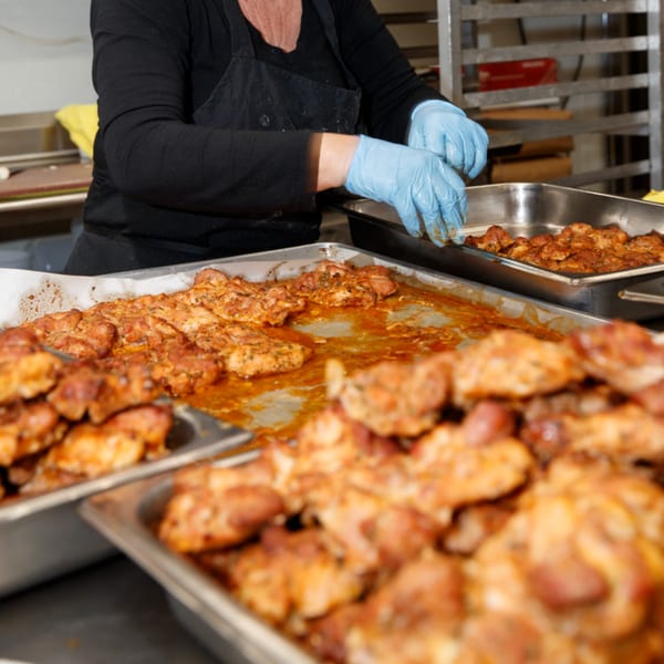 Gloved hands arrange seasoned chicken pieces in large metal trays at Cortina's Italian Market.