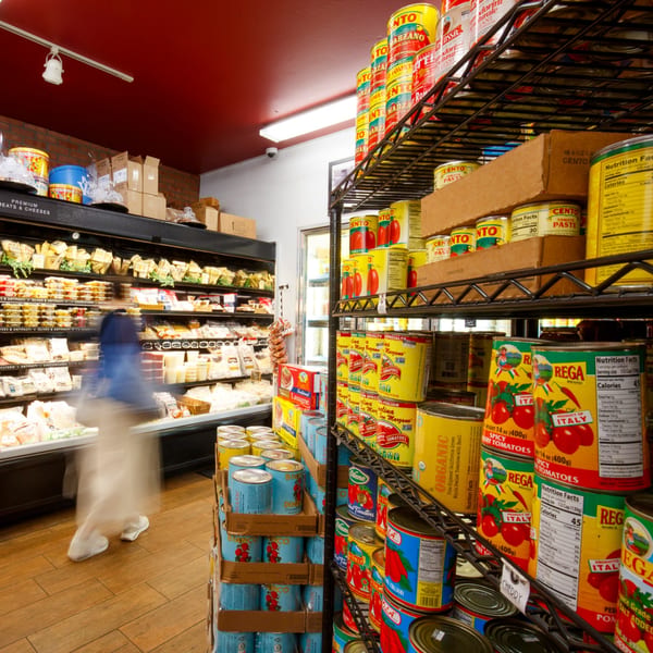 Aisle of an Italian market with shelves stocked with canned tomatoes and a deli case.