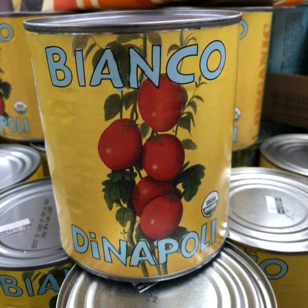 A yellow can of Bianco DiNapoli organic whole peeled tomatoes, with other cans stacked around it.