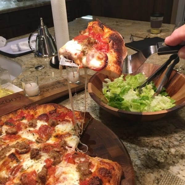 A hand lifts a slice of pepperoni and sausage pizza with melted cheese, next to a salad.
