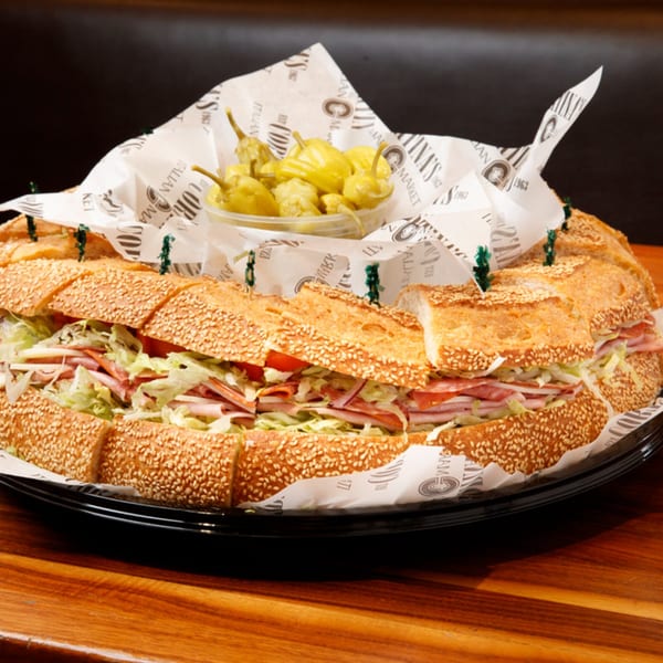 A large, circular Italian sub sandwich with deli meats, cheese, lettuce, and tomatoes, served with pickled peppers.