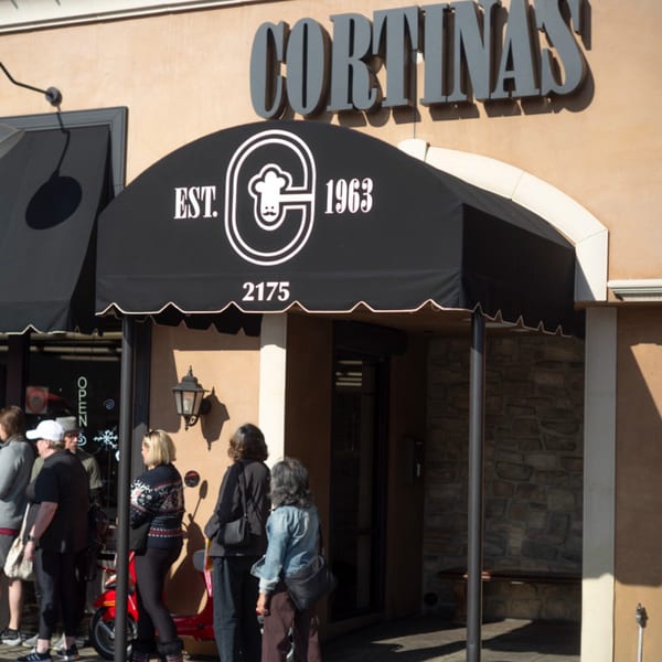 People queue outside Cortina's Italian Market & Pizzeria, with a black awning and sign visible.