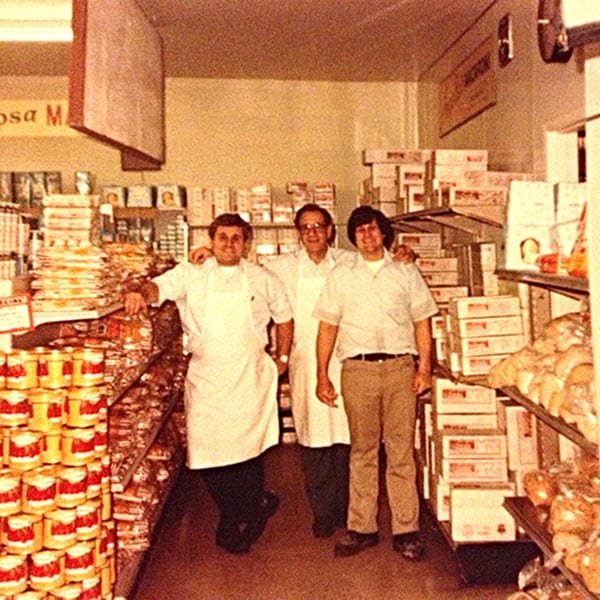 Three men, two wearing aprons, stand smiling in a vintage Italian market aisle.