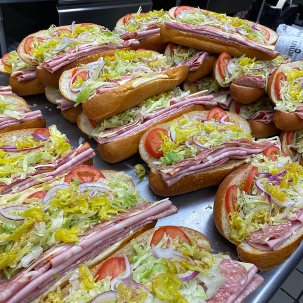 A large pile of Italian deli sandwiches, overflowing with meats, cheese, lettuce, tomatoes, and peppers.
