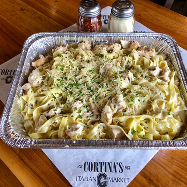 A large aluminum tray of Cortina's chicken alfredo pasta, topped with cheese and parsley.