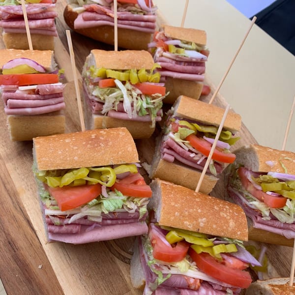 Many small Italian deli sandwiches with various meats, cheese, lettuce, tomatoes, and peppers on a wooden board.