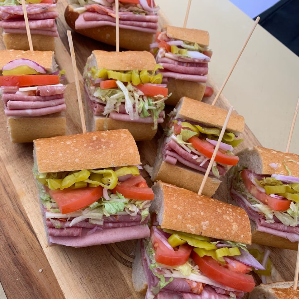 Many small Italian deli sandwiches with various meats, cheese, lettuce, tomatoes, and peppers on a wooden board.