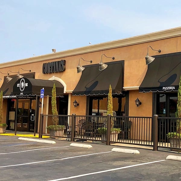 Exterior of Cortina's Italian Market & Pizzeria with black awnings, outdoor seating, and a parking lot.
