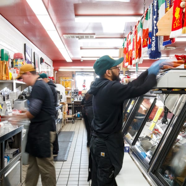 Workers in an Italian market, one stocking a deli case, with Italian flags and scarves overhead.
