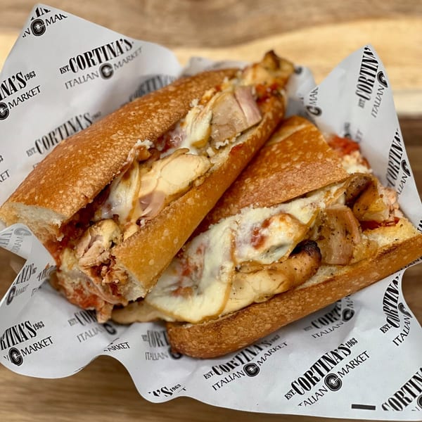 Two halves of a chicken parmesan sandwich with melted cheese and sauce, wrapped in Cortina's branded paper.