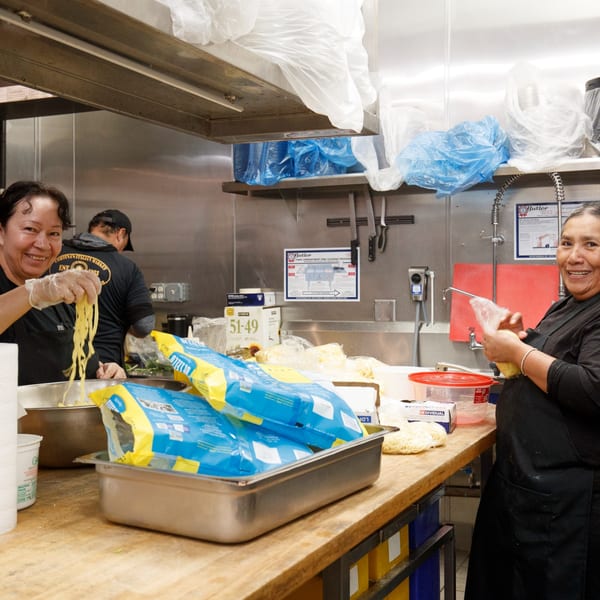 Two smiling Cortina's Italian Market employees preparing fresh pasta in a bustling kitchen.