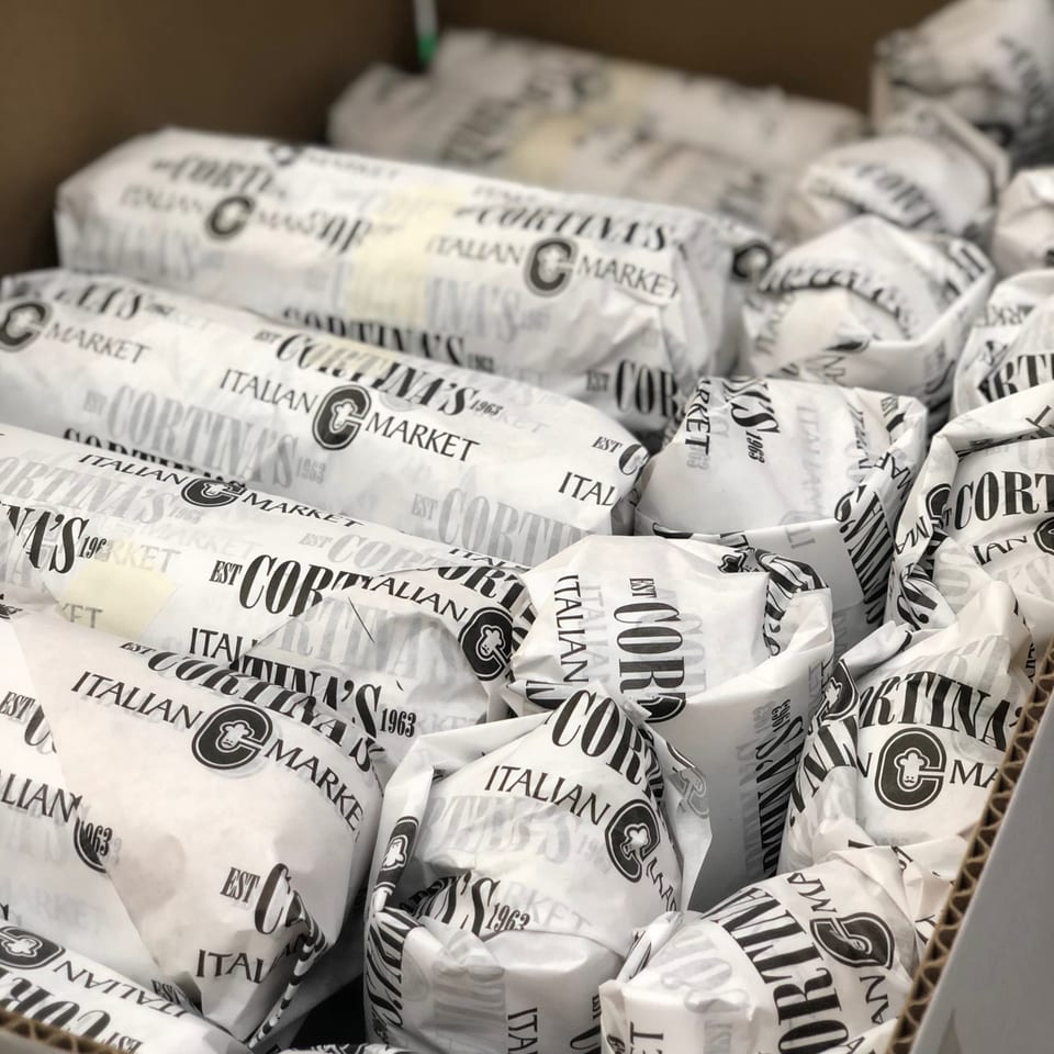 Cortina's Italian Market sandwiches, individually wrapped in branded paper, fill a cardboard box.