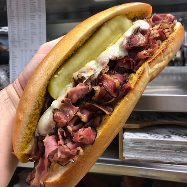 A hand holds a large pastrami sandwich with melted cheese, mustard, and a pickle spear.