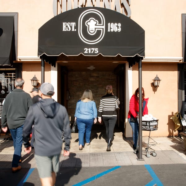 People entering Cortina's Italian Market & Pizzeria under a black awning with the restaurant's logo.