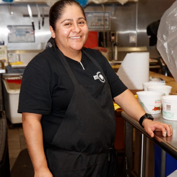 Smiling Cortina's Italian Market & Pizzeria employee in a black apron and shirt stands in the kitchen.