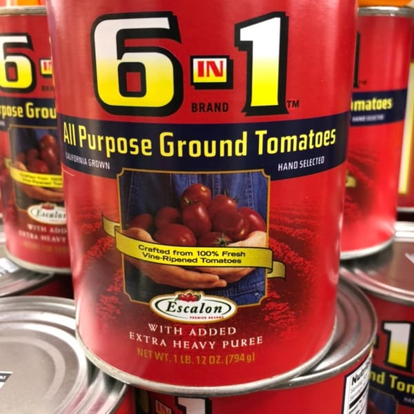 Close-up of a red can of Escalon 6-in-1 All Purpose Ground Tomatoes, with other cans blurred in the background.