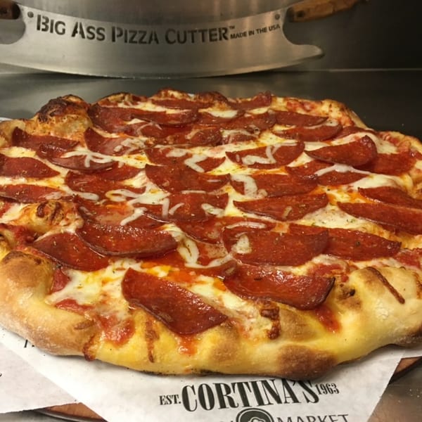 A pepperoni pizza from Cortina's Italian Market with a large pizza cutter in the background.